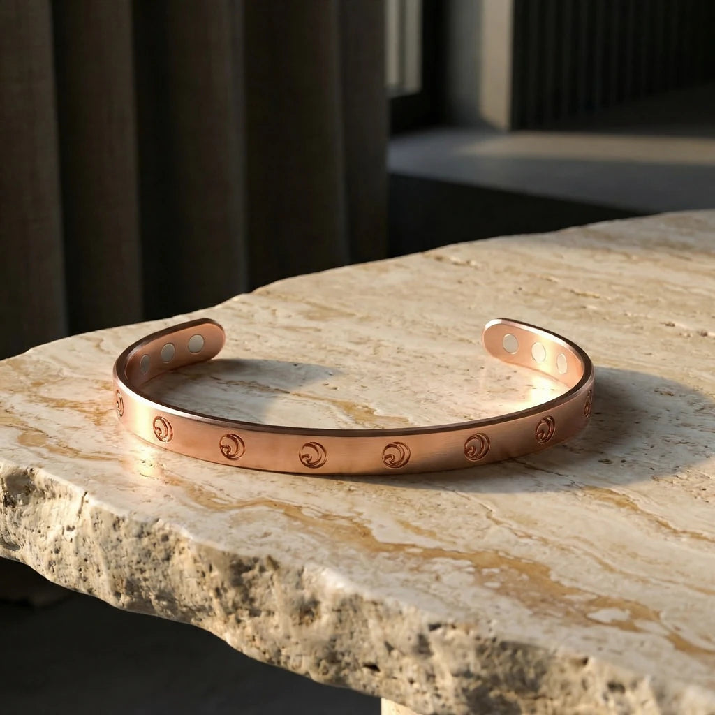A copper magnetic cuff bracelet with an engraved crescent moon pattern, resting on a stone surface.